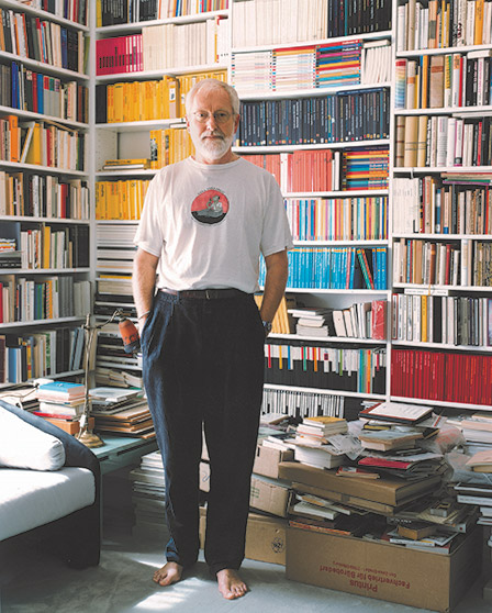 A man standing barefoot in a living room with numerous books in the background and on the floor