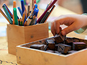 Eine Kinderhand greift nach einem Stempel auf einem Tisch mit Schreib- und Bastelmaterialien.
