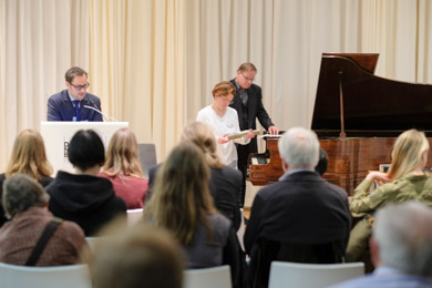 Das Publikum einer Veranstaltung schaut auf das Rednerpult an dem ein Mann vorträgt. Zugleich sind eine Frau und ein Mann an einem Reproduktionsflügel vor dem Publikum damit beschäftigt, eine Klavierrolle auszutauschen  Das Publikum einer Veranstaltung schaut auf das Rednerpult an dem ein Mann vorträgt. Zugleich sind eine Frau und ein Mann an einem Reproduktionsflügel vor dem Publikum damit beschäftigt, eine Klavierrolle auszutauschen